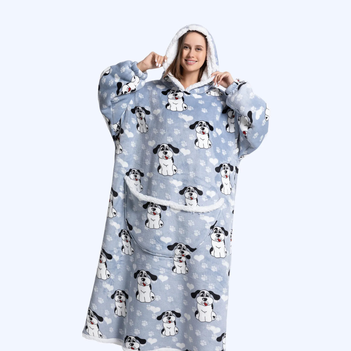 A woman wearing a light blue hoodie blanket with an all-over print of black-and-white dog faces and small white paw prints, posed against a plain light gray studio background.