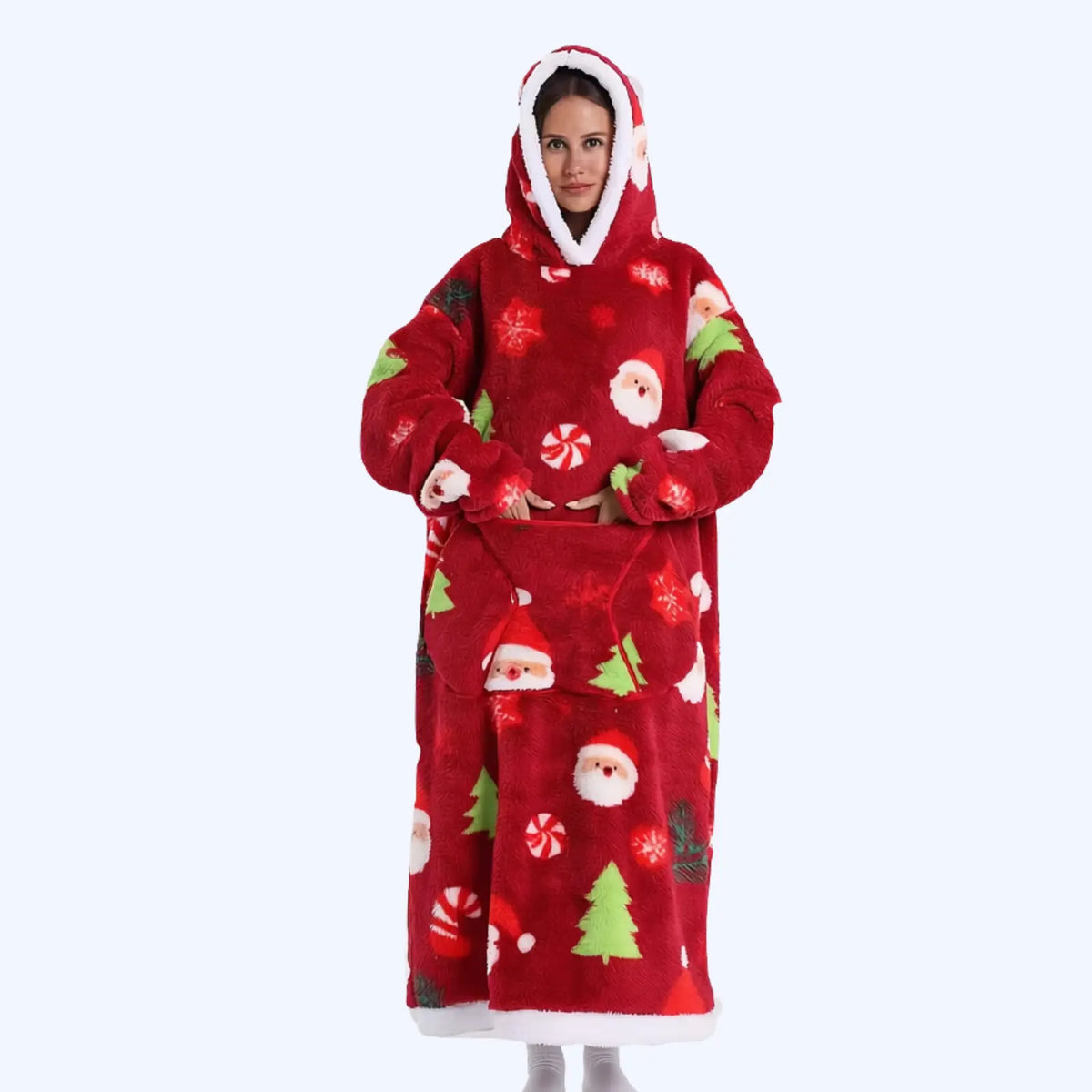 A person wearing a red plush hoodie blanket with a Christmas print featuring Santa Claus, candy canes, pine trees, and snowflakes, photographed against a plain light gray background.