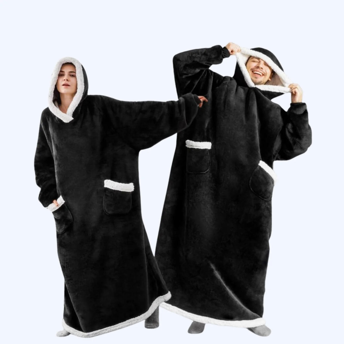 A man and a woman wearing matching black hoodie blankets with white faux-fur trim, standing against a light gray studio background.