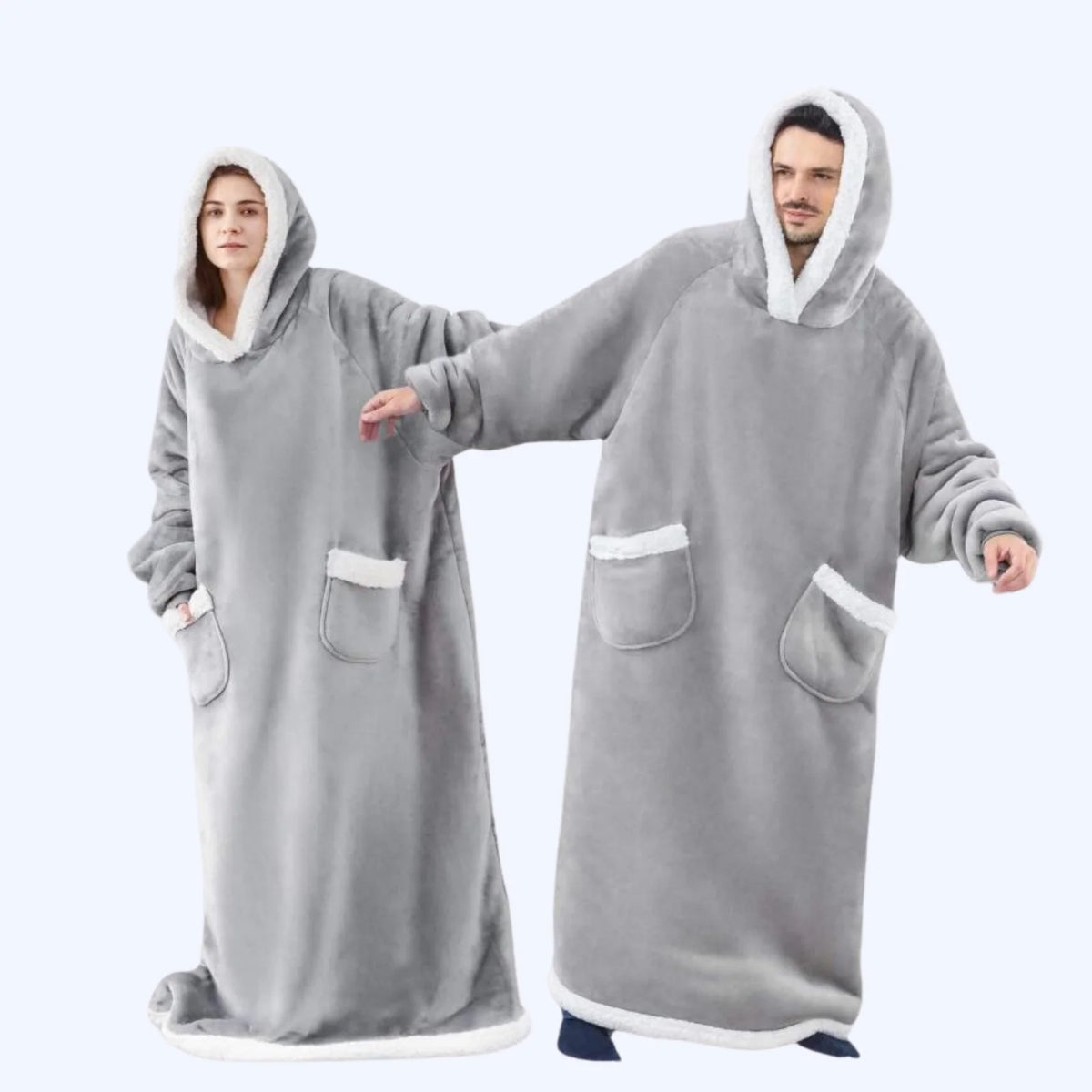 A man and a woman wearing matching light grey hoodie blankets with white faux-fur trim, standing against a light gray studio background.