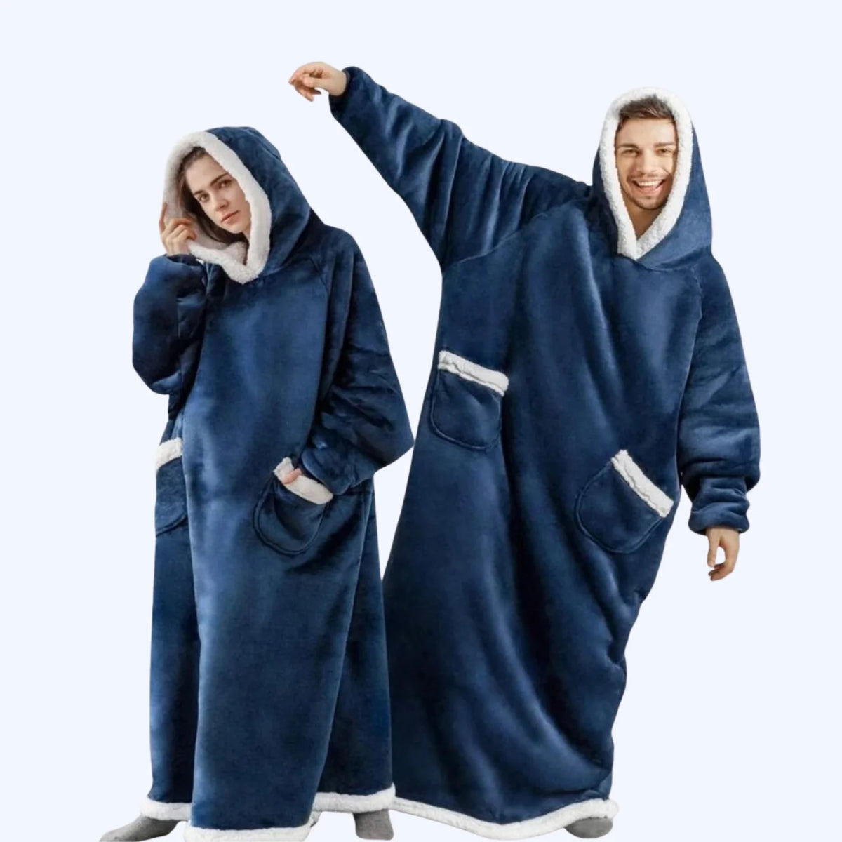 A man and a woman wearing matching navy blue hoodie blankets with white faux-fur trim, standing against a light gray studio background.