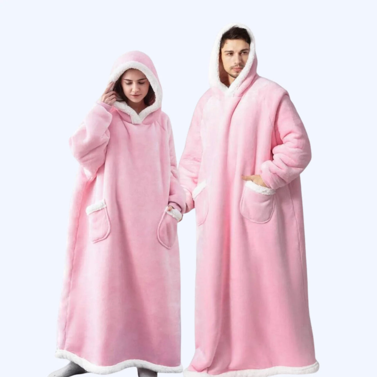 A man and a woman wearing matching pink hoodie blankets with white faux-fur trim, standing against a light gray studio background.