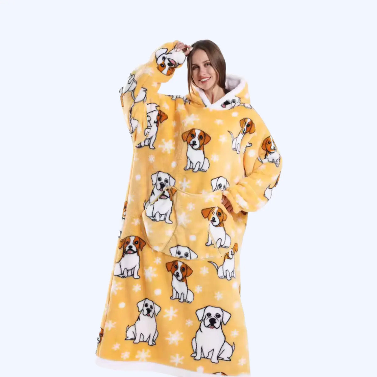 A woman wearing a yellow hoodie blanket printed with Jack Russell Terrier dogs and white snowflakes, set against a plain light blue background.