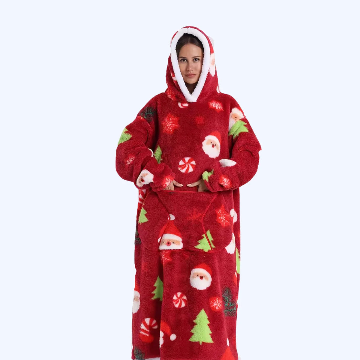 A person wearing a red plush hoodie blanket with a Christmas print featuring Santa Claus, candy canes, pine trees, and snowflakes, photographed against a plain light gray background.