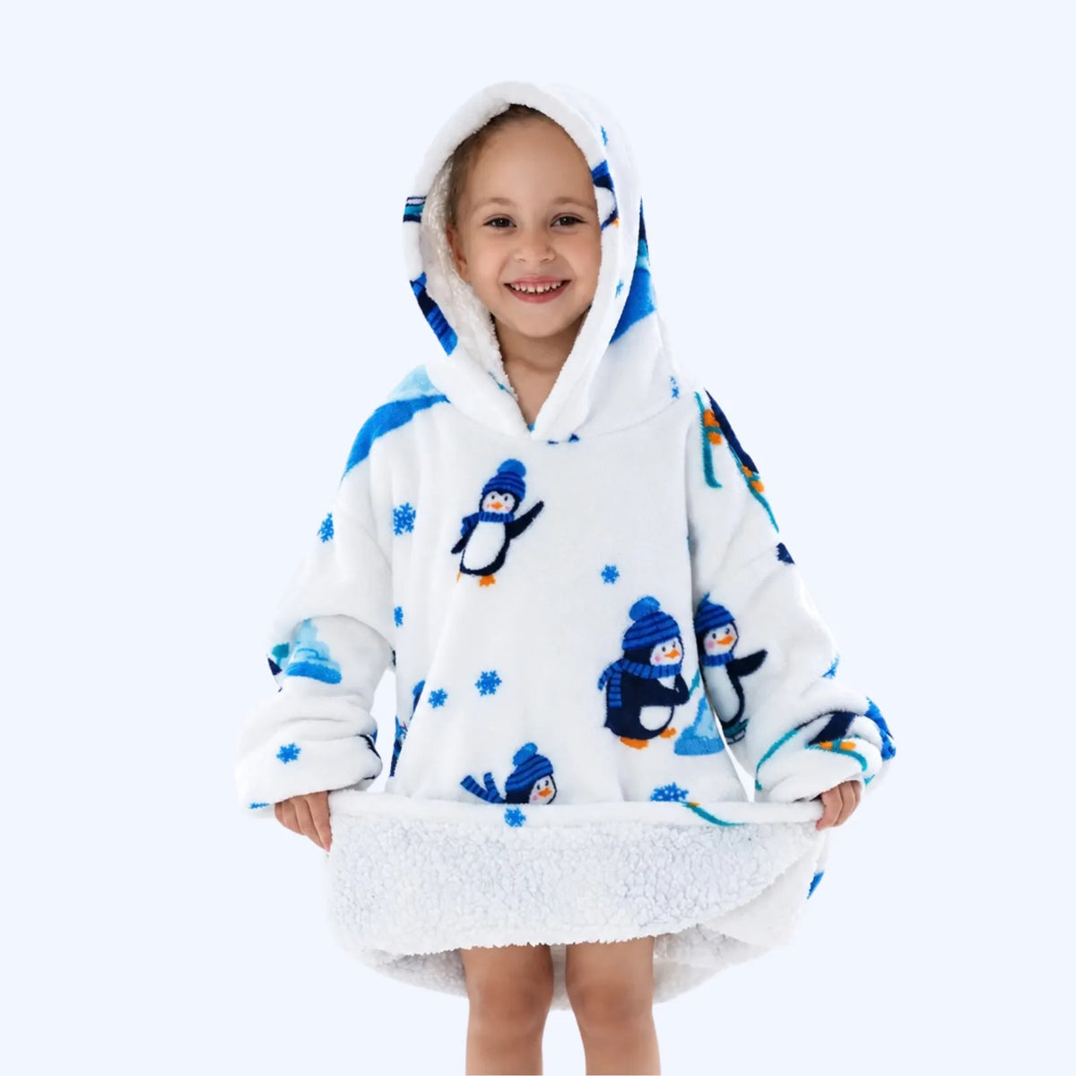 A young child wearing a white hoodie blanket with blue penguin prints and blue snowflake details, standing against a light gray studio background.